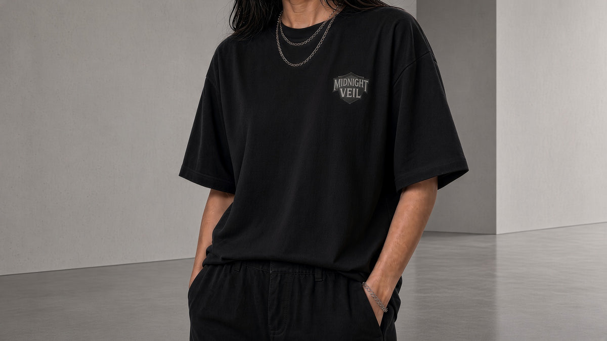 Woman wearing an oversized Obsidian Black Core Emblem box tee with Midnight Veil chest shield, hands in pockets, studio concrete background